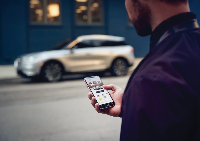 A man is holding his phone looking at the Lincoln Way™ App and we see his 2022 Lincoln Corsair SUV in the background against a blue brick wall | Irwin Lincoln in Freehold NJ
