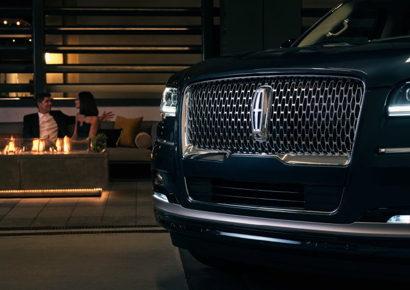 The signature Lincoln grille on the 2022 Lincoln Navigator SUV commands attention as a couple relaxes by a fire beyond | Irwin Lincoln in Freehold NJ