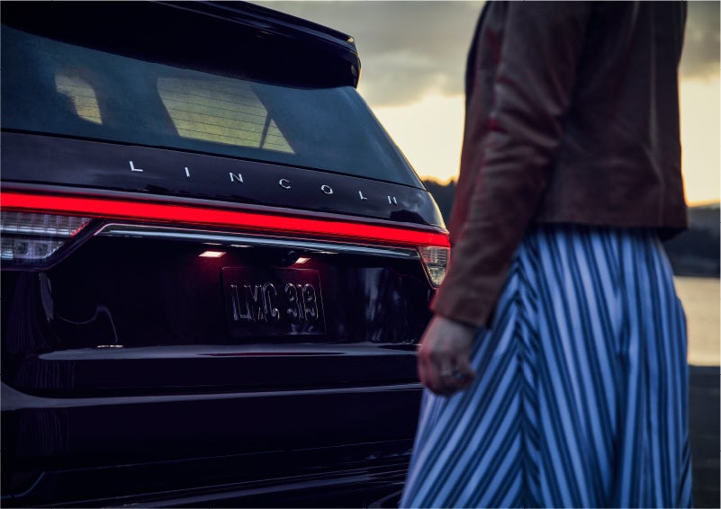 A person is shown near the rear of a 2023 Lincoln Aviator® SUV as the Lincoln Embrace illuminates the rear lights | Irwin Lincoln in Freehold NJ